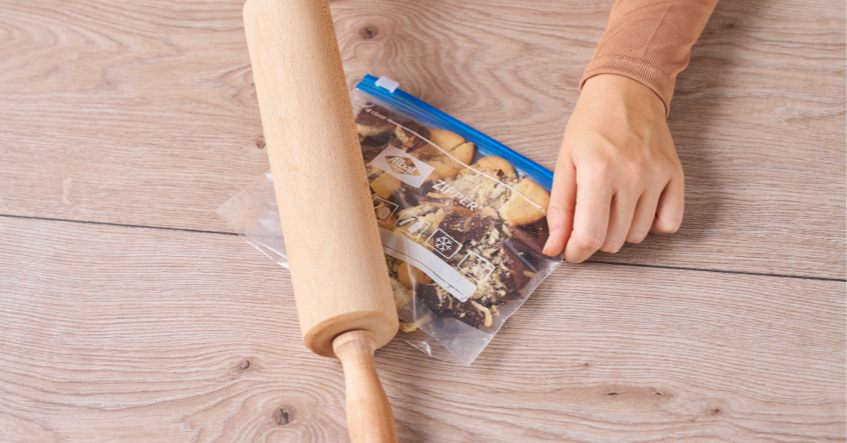 Eine Hand hält einen Toppits Zipper Gefrierbeutel mit Keksen auf einem Holztisch fest, während ein Nudelholz darüber rollt, um die Kekse zu zerkleinern
